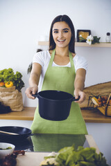 An attractive young dark-haired woman offering ready soup by new keto recipe while standing and smiling in the kitchen. Cooking and householding concepts