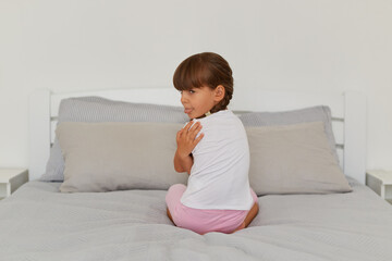 Indoor shot of offended female kid sitting on the bed, turning to camera and showing tongue out,...