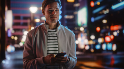 Handsome Man Using Smartphone Walking Through Night City Full of Neon Colors and Entertainment....