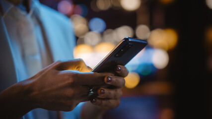Focus on Female Hand Using Smartphone Walking Through Night City Street Full of Neon Light. Woman Holding Mobile Phone, Posting Social Media, Online Shopping, Texting.
