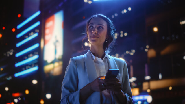 Beautiful Young Woman Using Smartphone Standing on the Night City Street Full of Neon Light. Portrait of Gorgeous Smiling Female Using Mobile Phone.