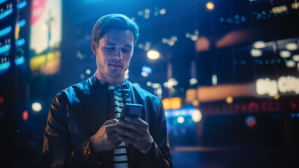 Man Using Smartphone Walking Through Night City Street Full of Neon Light. Smiling Stylish Man Using Mobile Phone, Social Media, Online Shopping, Texting on Dating App.