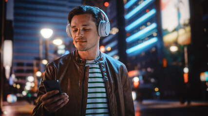Portrait of Handsome Man Wearing Headphones Walking Through Night City Street Full of Neon Light....
