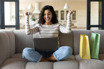 Excited young woman with laptop and shopping bags making YES gesture, buying things online from home