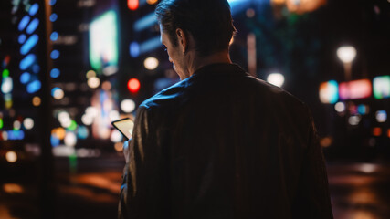 Following Shot of Handsome Man Using Smartphone Walking in a Modern City Street with Neon Lights at...