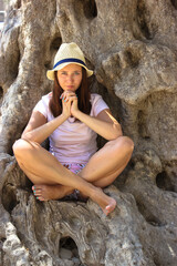 A serious beautiful young woman 20-30 years old wearing a straw hat sitting under an ancient olive tree in a lotus pose and thinking. A tourist girl on summer vacation in Greece. Unity with nature.