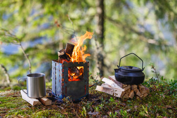 A burning camping wood stove with a kettle, a mug and firewood on the background of the forest....