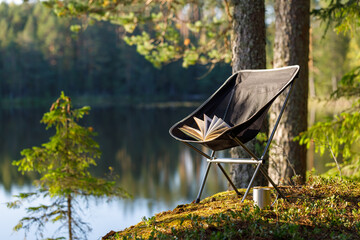 Camping folding chair with a book and a mug on the background of a forest lake. The chair is in focus, the background is blurred. - obrazy, fototapety, plakaty