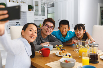 Smiling asian parents with son and daughter sitting at breakfast table taking selfie