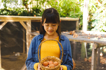 Asian girl smiling and holding basket, collecting eggs from hen house in garden © wavebreak3