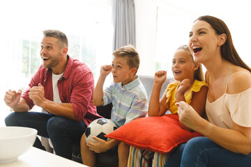 Excited caucasian parents on couch with daughter and son watching football match on tv and cheering