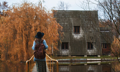 Traveler woman in hat with backpack walking across the bridge to old wooden house, touristic place, beautiful fall season