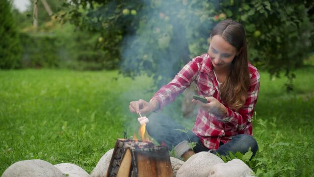 Young woman using a smartphone while sitting by a campfire on a picnic