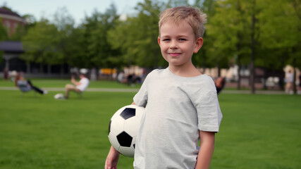 Happy blonde preschool boy standing in football field with soccer ball looking at camera
