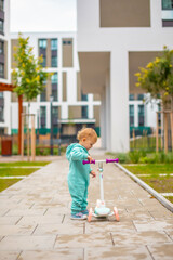 Cute little toddler girl in blue overalls riding on kick scooter. Happy healthy lovely baby child having fun in the city. Active kid on cold day outdoors.