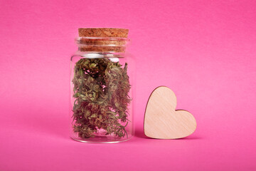 marijuana love symbol, jar with dry cannabis buds and a wooden heart on pink background.