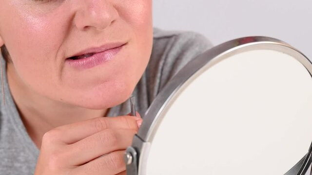 Caucasian woman looks in the mirror and removes hair on her chin herself with the help of tweezers. Hirsutism.