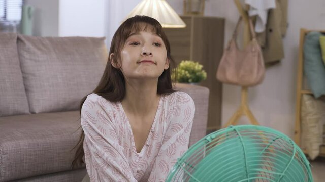 happy asian female is using an electric fan to escape high temperature and leaning close to it while it is blowing on her face in a hot summer night at home.