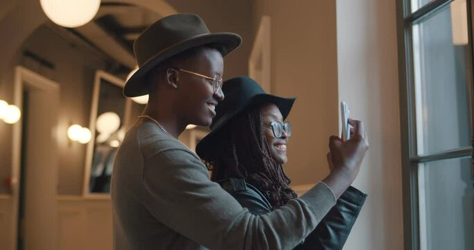 Smiling afro-american couple standing near window taking photo on smartphone