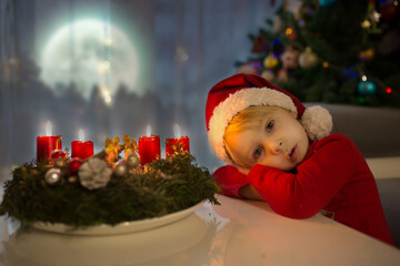 Beautiful toddler child, boy, waiting on the window on Christmas eve, looking for Santa Claus