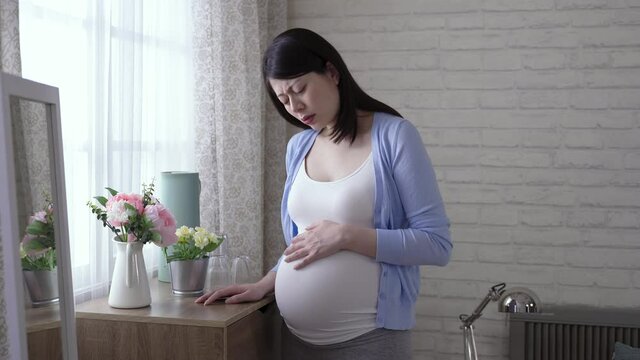 asian expectant female standing by the cupboard is holding her tummy and groaning in pain due to labor contraction at bright home interior.