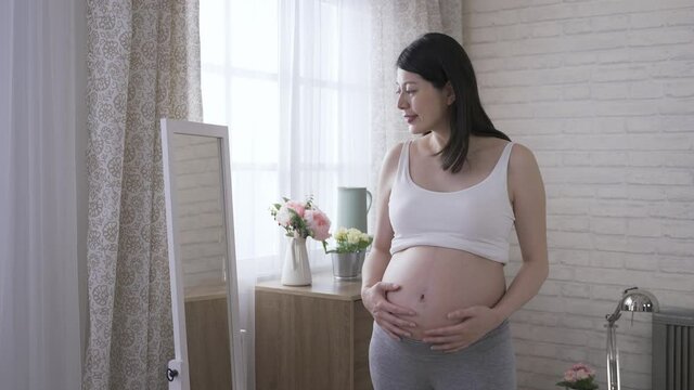 happy expectant woman is looking at herself in the mirror and feeling her tummy with anticipation in the morning at home.