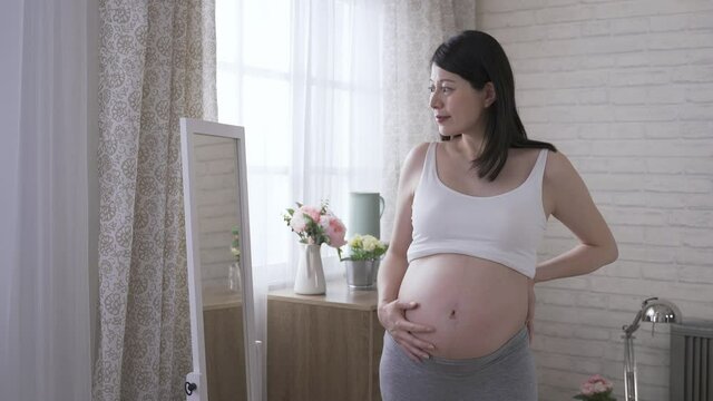 pretty pregnant woman standing by the mirror with hands on lower back is touching her tummy softly while staring at her reflection with a smile.