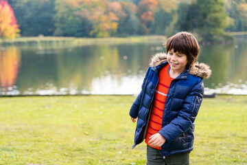 Young boy enjoying a walk in the autumn park near beautiful lake with reflections. Child wearing warm jacket smiling and playing outdoors. Golden fall weather. © Anna