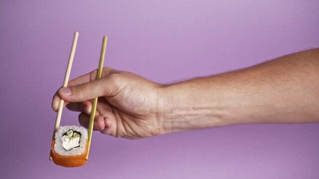Close up on male caucasian hand holding wooden chop sticks with one roll sushi, isolated on puple background. Sushi restaurant, food delivery,  asian food, japanese cuisine delivery. 