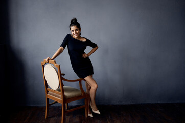 Fototapeta na wymiar brunette in a black dress near the chair luxury fashion dark background