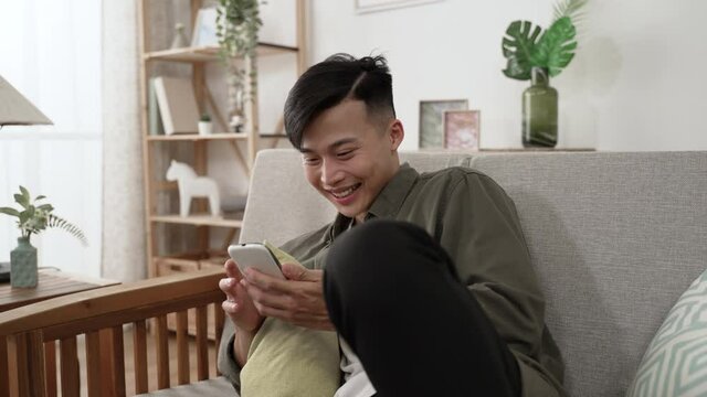 Carefree Asian Young Man Throwing Head Back And Laughing Loudly While Watching A Funny Video On His Mobile Phone On Comfortable Sofa In The Living Room At Home.