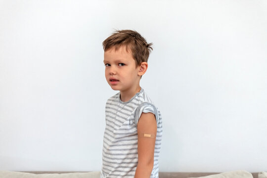 Sad Caucasian boy showing adhesive bandage on shoulder after getting vaccinated during COVID-19 pandemic isolated on the white background. Child showing his arm with bandage after getting vaccine.