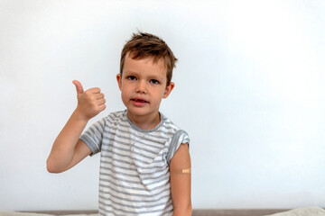 Cute little boy with adhesive bandage plaster on his arm after vaccination. Injection covid vaccine, healthcare for children and teenagers. Vaccinated boy showing arm after vaccination with thumb up.