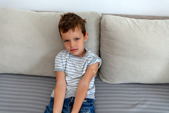 Unhappy cute little boy sitting against a neutral background after receiving Coronavirus vaccine, looking at camera. Crying Caucasian 6 years old boy after being vaccinated wearing a band aid on arm.