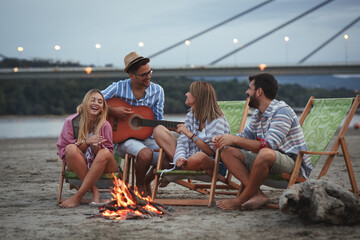 On a tranquil autumn evening, a group of friends gathers by the riverside sandy beach. They sit around a small campfire, under the starlit sky, enjoying their drinks, singing, and playing guitar.	