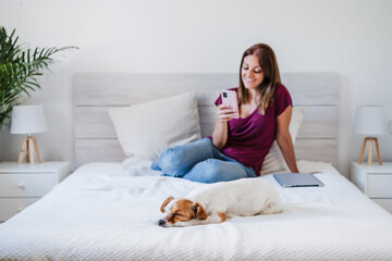 daydreaming caucasian woman at home working on laptop and mobile phone while cute jack russell dog resting on bed. Home office, Pets, love and relax. Selective focus on dog