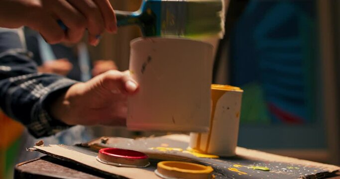 Shot of a can of paint standing on a table by an easel, a dirty paintbrush lies on top of it, female hands grab objects and hand them to a friend painting abstract lines in solid color on a canvas
