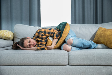 Young woman with menstruation is lying down on the couch and holding on to her stomach because she...