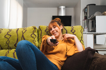 Obraz premium Caucasian female in an orange shirt changing the channel with a remote