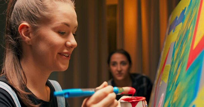 Smiling brunette with hair tied up in ponytail is standing in front of canvas on which she is painting, using red paint and professional brush, telling friend story about beginning of artistic career