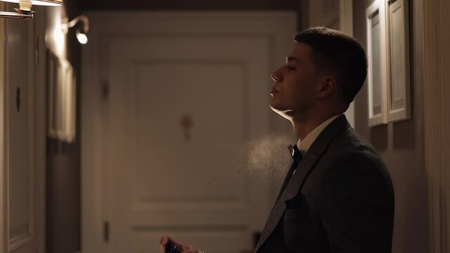 Groom splits the perfume or deodorant on hisself. Wedding day. Slow motion. Well dressed young adult handsome businessman using perfume from a bottle on his neck in dark room background. Backlit