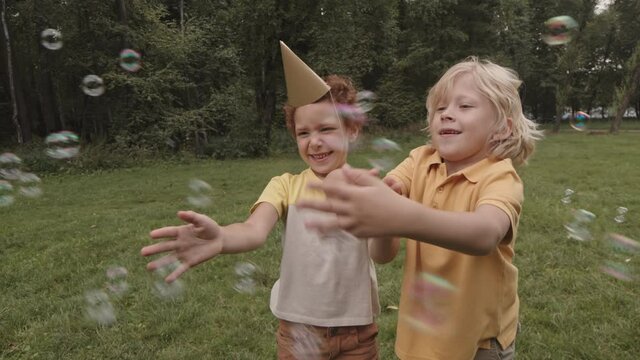 Medium of two multiethnic five-year-old boys smiling, playing with soap bubbles catching them in summer park on lawn, kid wearing party hat