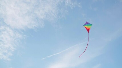 4K multicoloured kite flying in blue sky under gusts of wind with some white clouds up high. Hobbies and leisure activities concept.