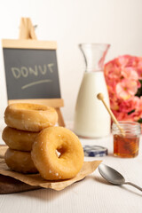 View of donuts on brown paper and white table with spoon, honey, milk bottle and blackboard, selective focus, white background, vertical