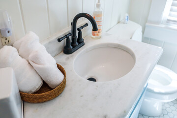 A basket of organized clean rolled white towels on a bathroom counter in a guest bathroom near a sink and toilet.