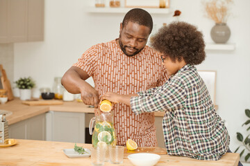 Cute little boy squeezing lemon juice into jug with lemonade while helping his father to make...