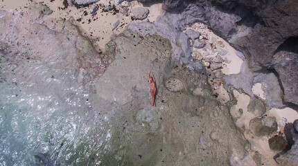 Young and beautiful fashion female model in bikini lying on the rock in the sea. Fashion shooting. Aerial video in Bali island. Relaxing on vacation