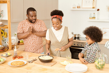 Young African female putting frying-pan with pasta on table among her husband and son