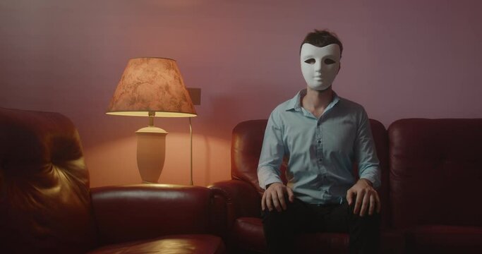 Ragazzo con maschera bianca anonima seduto sul divano saluta con la mano