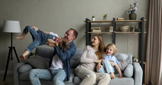 Cheery father sit on sofa lifts upside down cute 4s daughter, pregnant mother hugs son, couple and kids enjoy carefree weekend together at home. Bank mortgage, happy multi-member large family portrait
